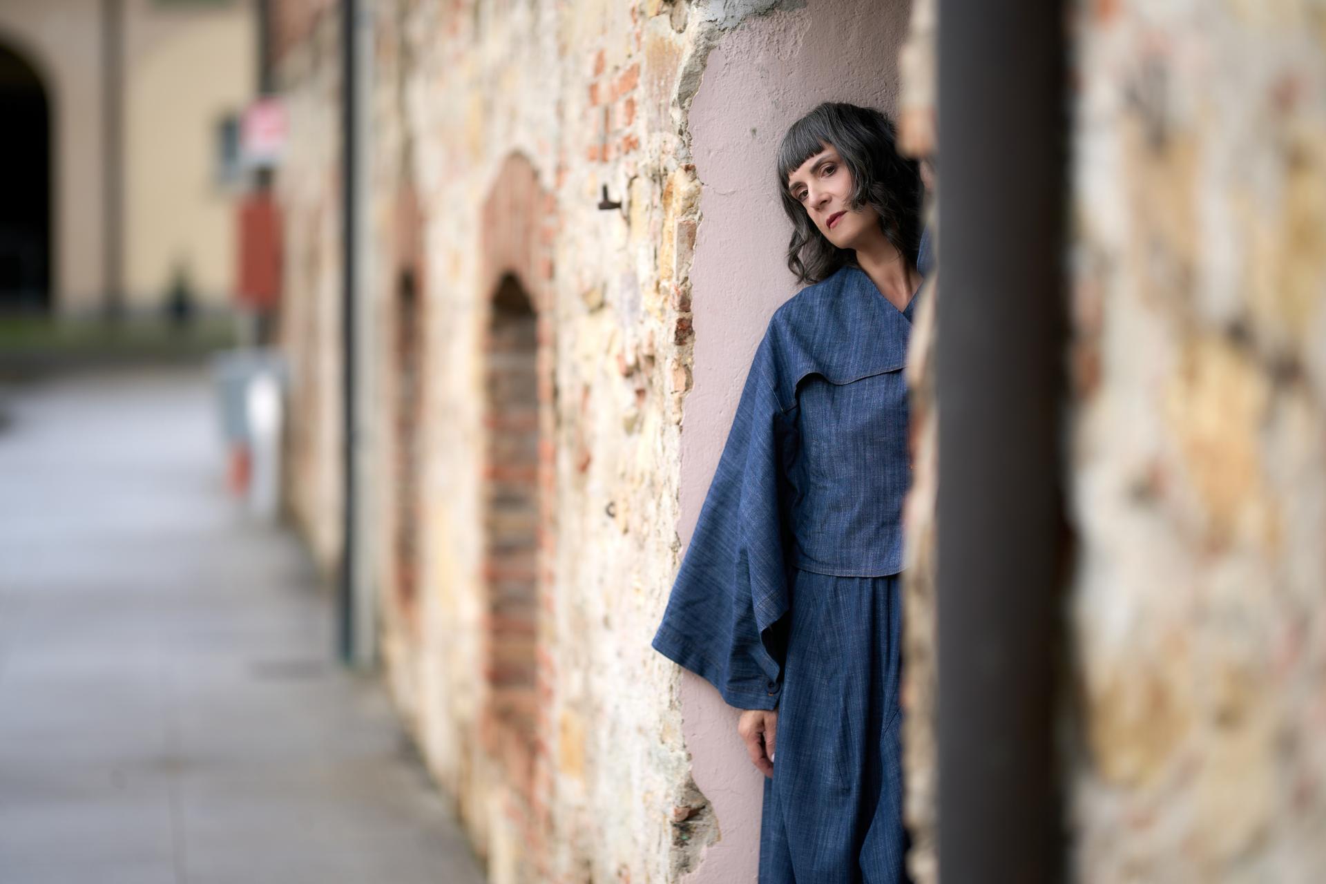 Una donna con capelli scuri e abito blu si appoggia a un muro di mattoni in un vicolo.