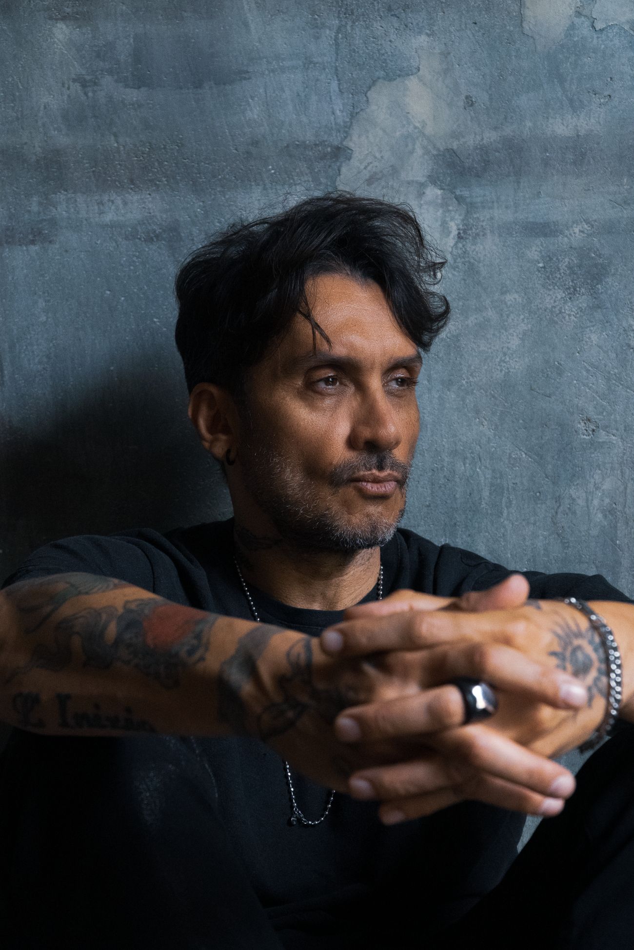 Ritratto di Fabrizio Moro con le mani intrecciate e lo sguardo pensieroso davanti a un muro grigio.